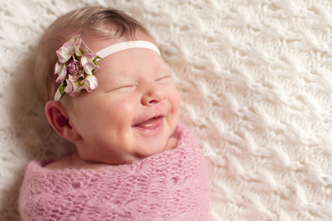Newborn baby laying on a white blanket wearing a pink wrap and a flower headband smiling for newborn photography