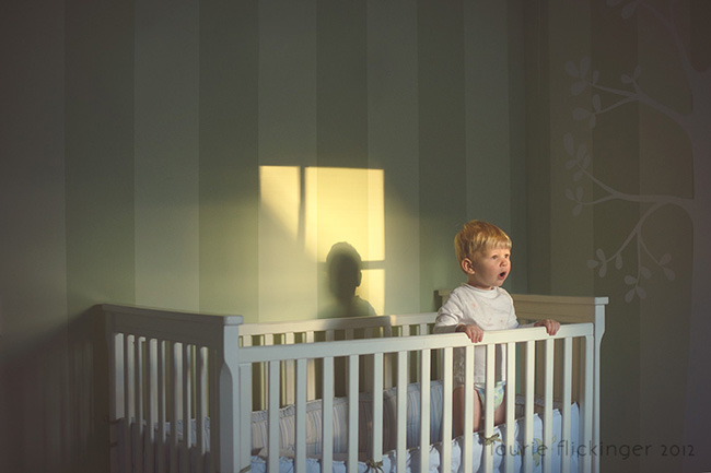 Child standing in a white crib looking out towards a window that you see the shadow casted on the wall. The image shows proper framing photography.