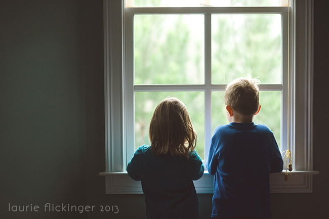Two children standing in front of a square window looking outside. The children are framed in the photograph  by the window itself.