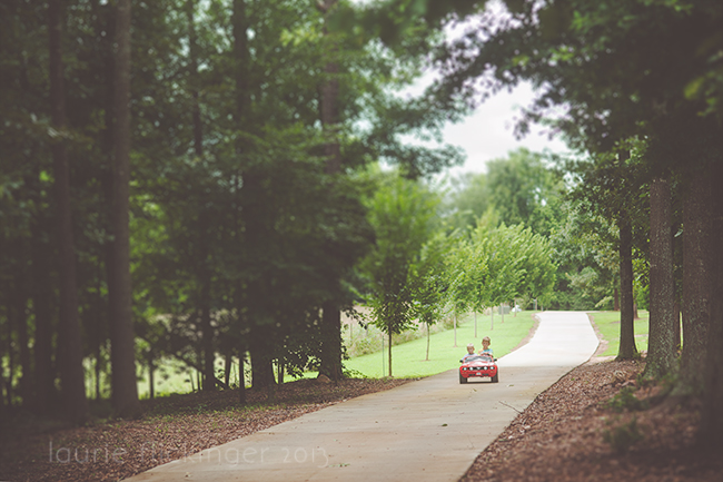 Two children driving toy car down a path using framing in photography with trees on both sides.