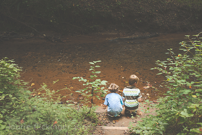 2 children framed by plants on both sides of them sitting next to a brown river.
