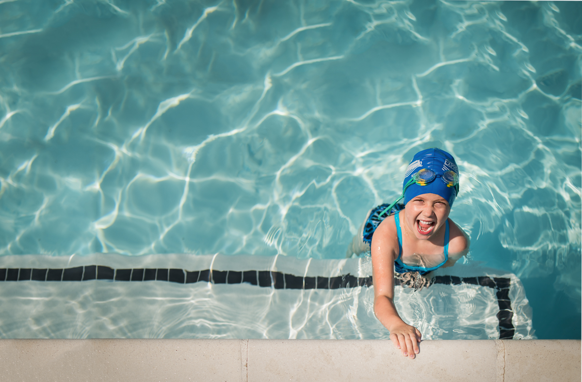 Child in a pool wearing a swim cap and goggles looking up at the photographer. Showing a tip of how to shoot in hard light without shadows.