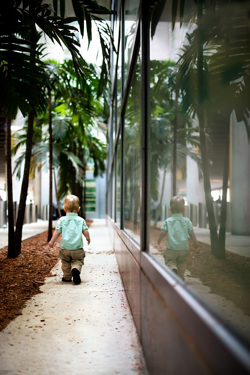 Child wearing a blue shirt with palm trees all around in the reflection photography in the window. 