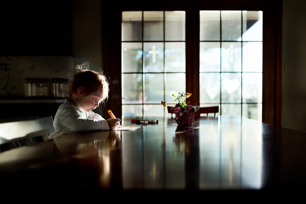 A child sitting at a table with a bright window filled door in the background with a reflection of the child for reflective photography.