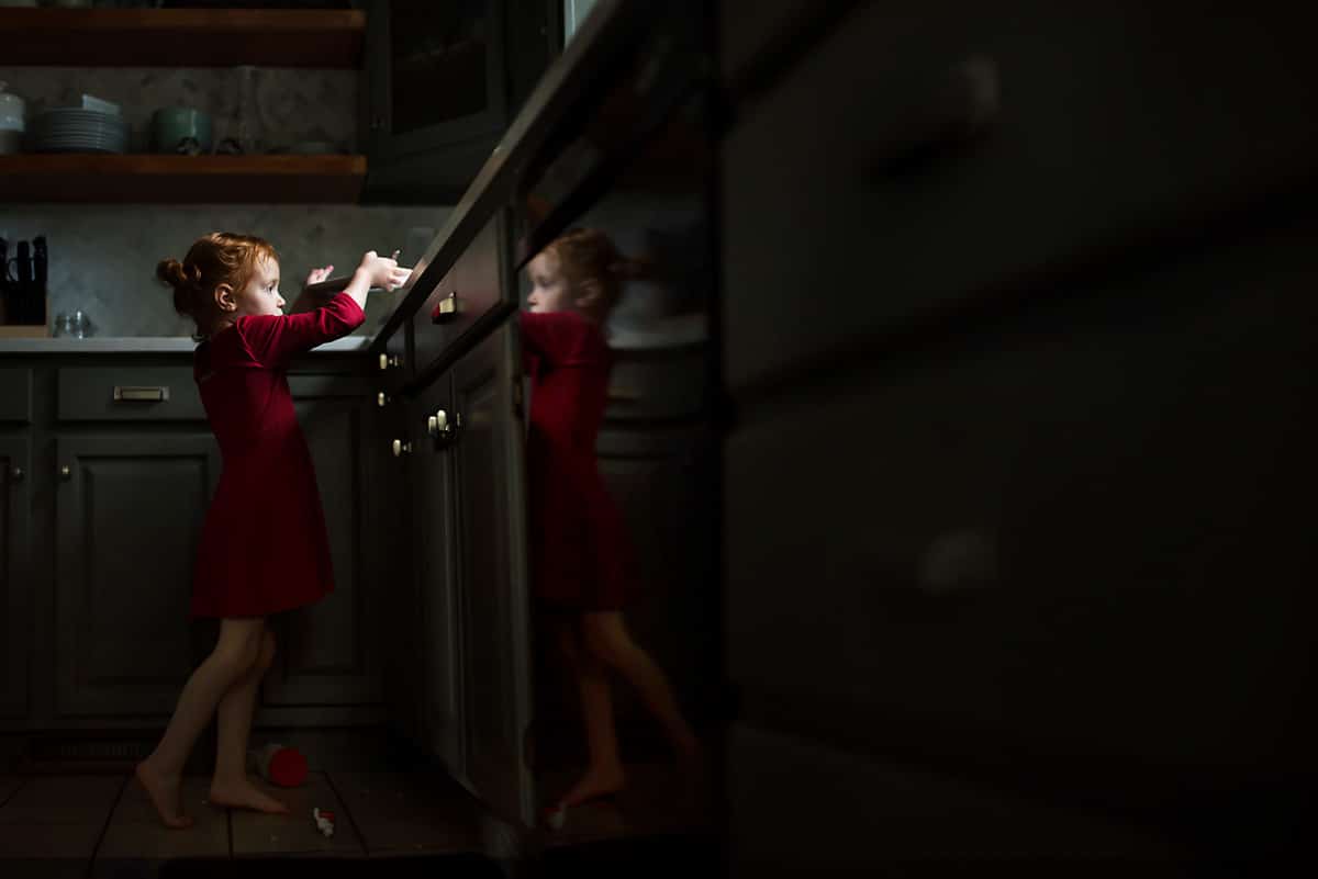 A child wearing a red dress in the kitchen with a reflection in the dishwasher. 