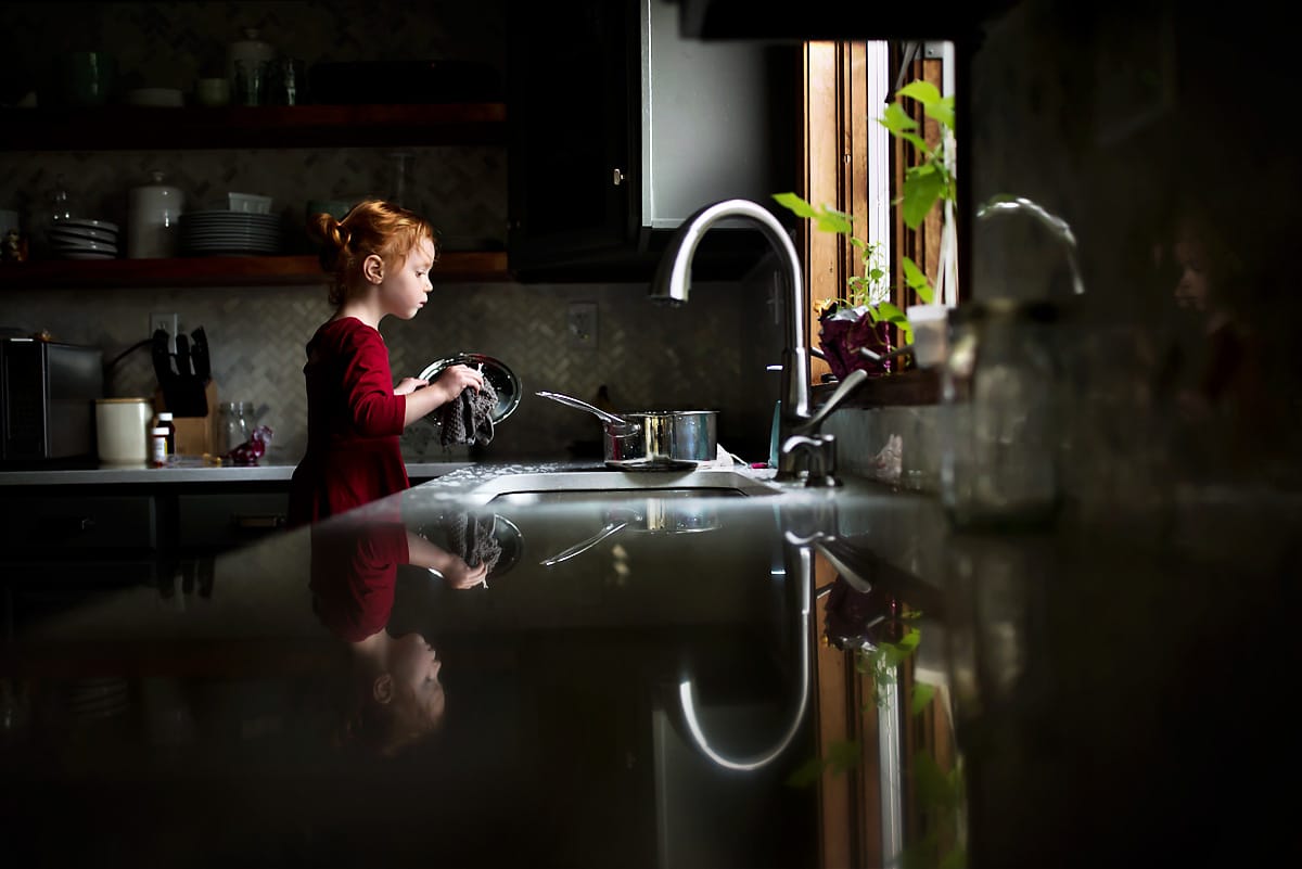 Child in a red dress washing dishes in the kitchen with a reflection on the counter top. This is an example of reflection photography.