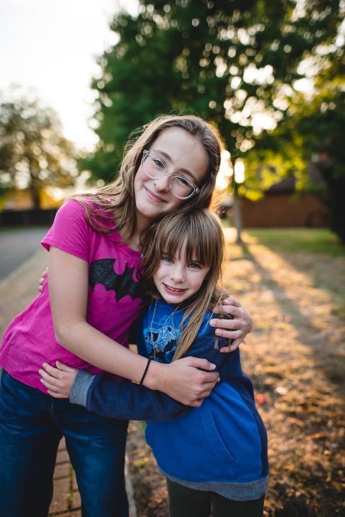 Two children hugging one is a pink shirt, one in a blue shirt. Nice bokeh coming in through the trees in the background. Backlighting photography during golden hour.