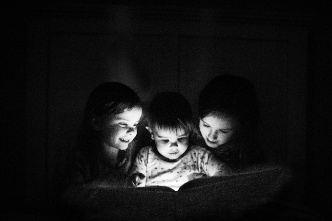 A black and white photo of three children reading a book in the dark with a flashlight. 