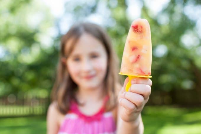 Child in pink shirt holding a fruit popsicle with a yellow handle. 
