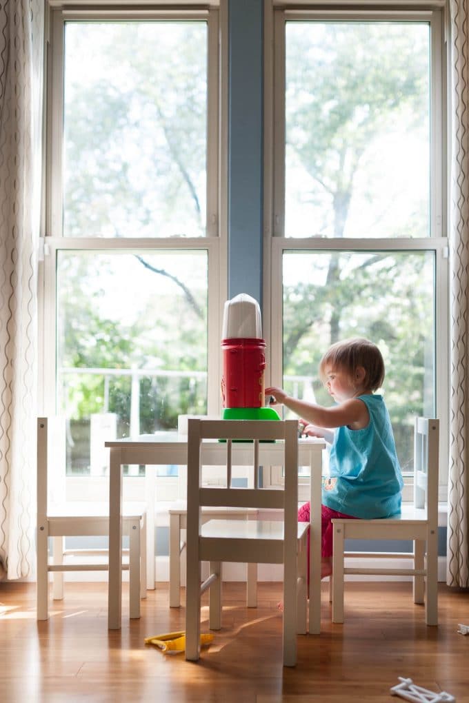Child playing in front of a big window sitting at a table with a red barn.