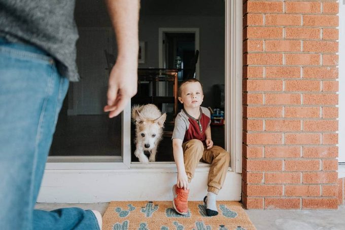 Child in a red shirt putting on shoes while a dog is running out of the door as an example of lifestyle photography tips